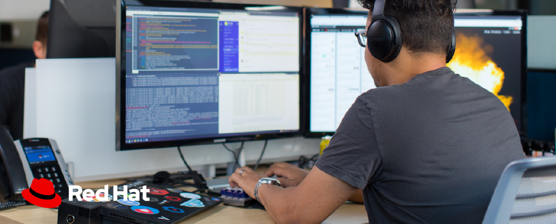 Photograph of a man in an office using a computer with the Red Hat logo overlayed on a busy part of the image. The logo is difficult to see.