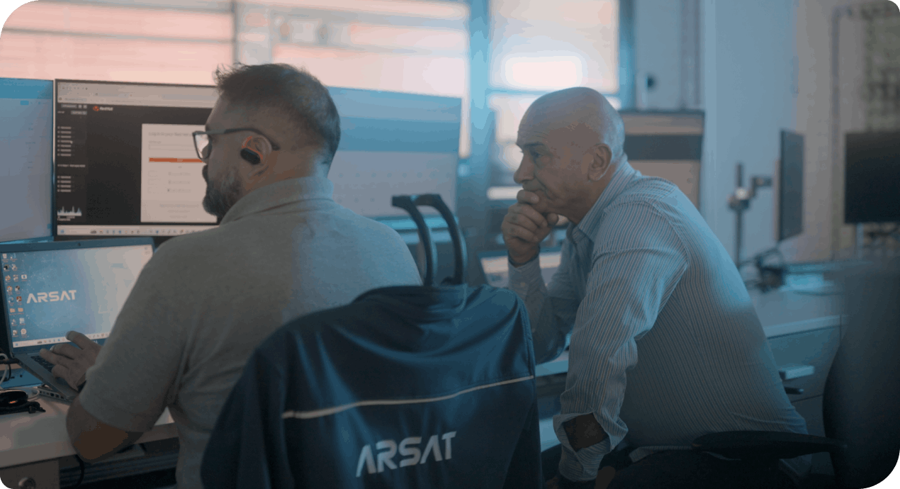 Two colleagues in a control room collaborate, one seated at a monitor and the other looking on thoughtfully
