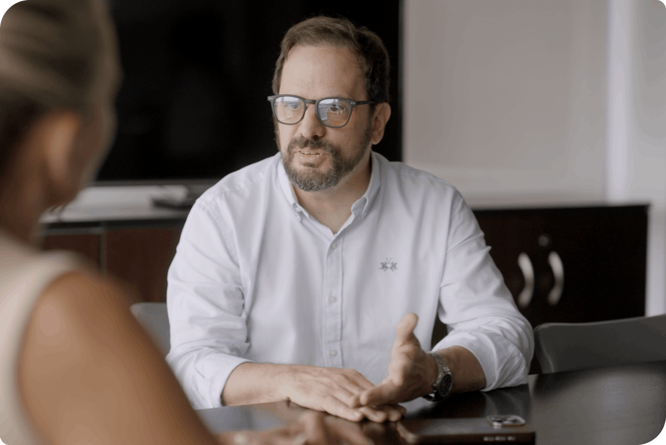 A man with glasses gestures as he speaks to a someone across a conference table