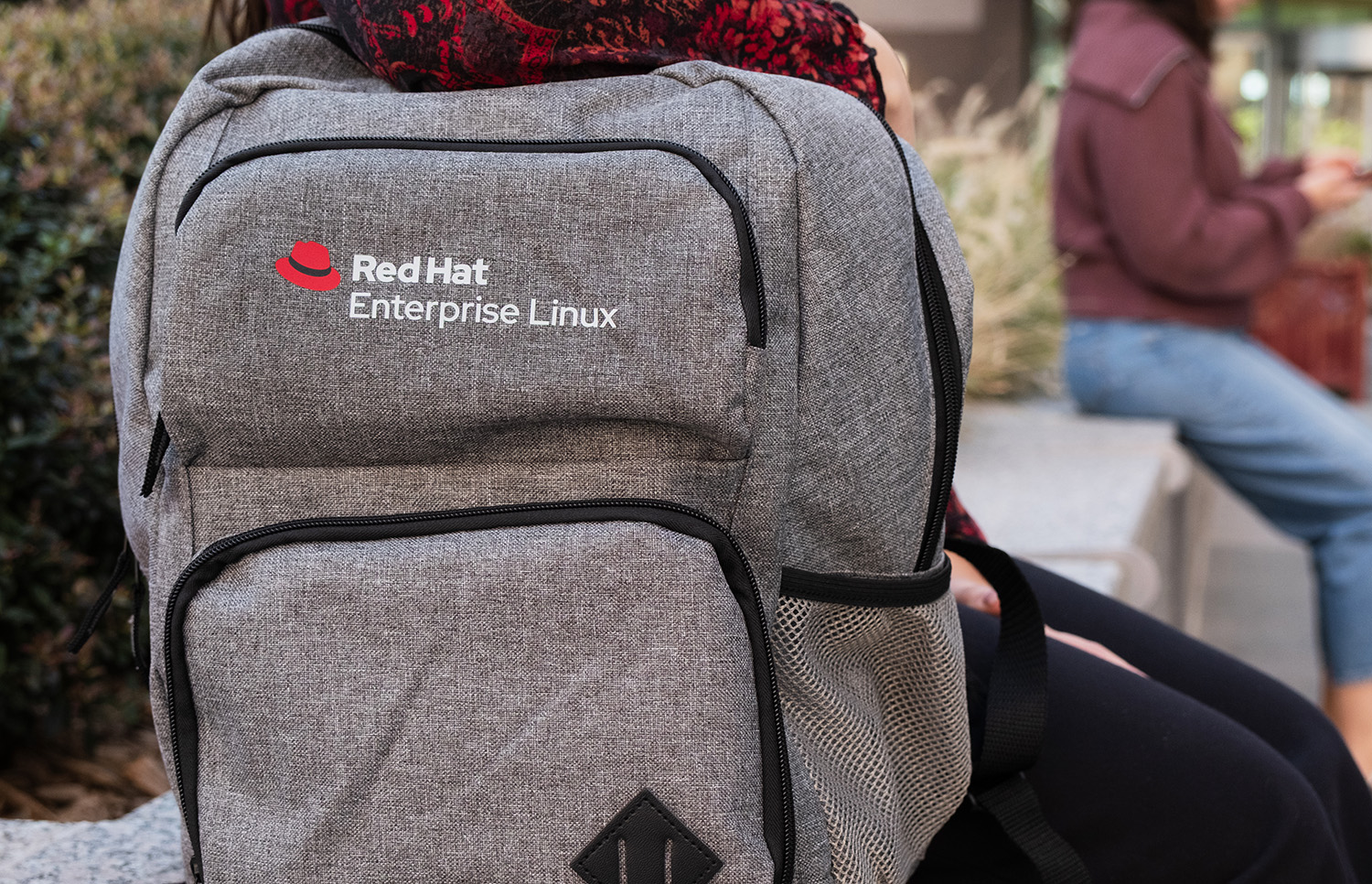 A woman sits with a gray backpack beside her. The Red Hat Enterprise Linux logo is printed in full color on the upper pocket.