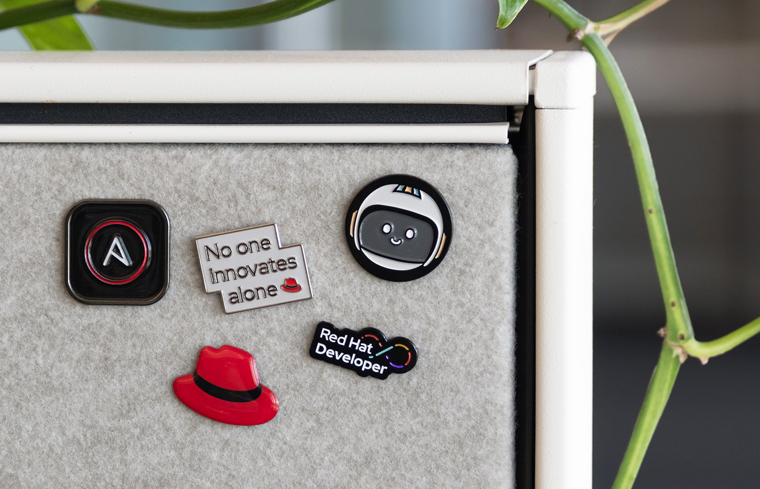 Red Hat enamel pins, including a red fedora, Repo the Red Hat Developer mascot, and the Red Hat Ansible icon, on a grey cubicle wall.
