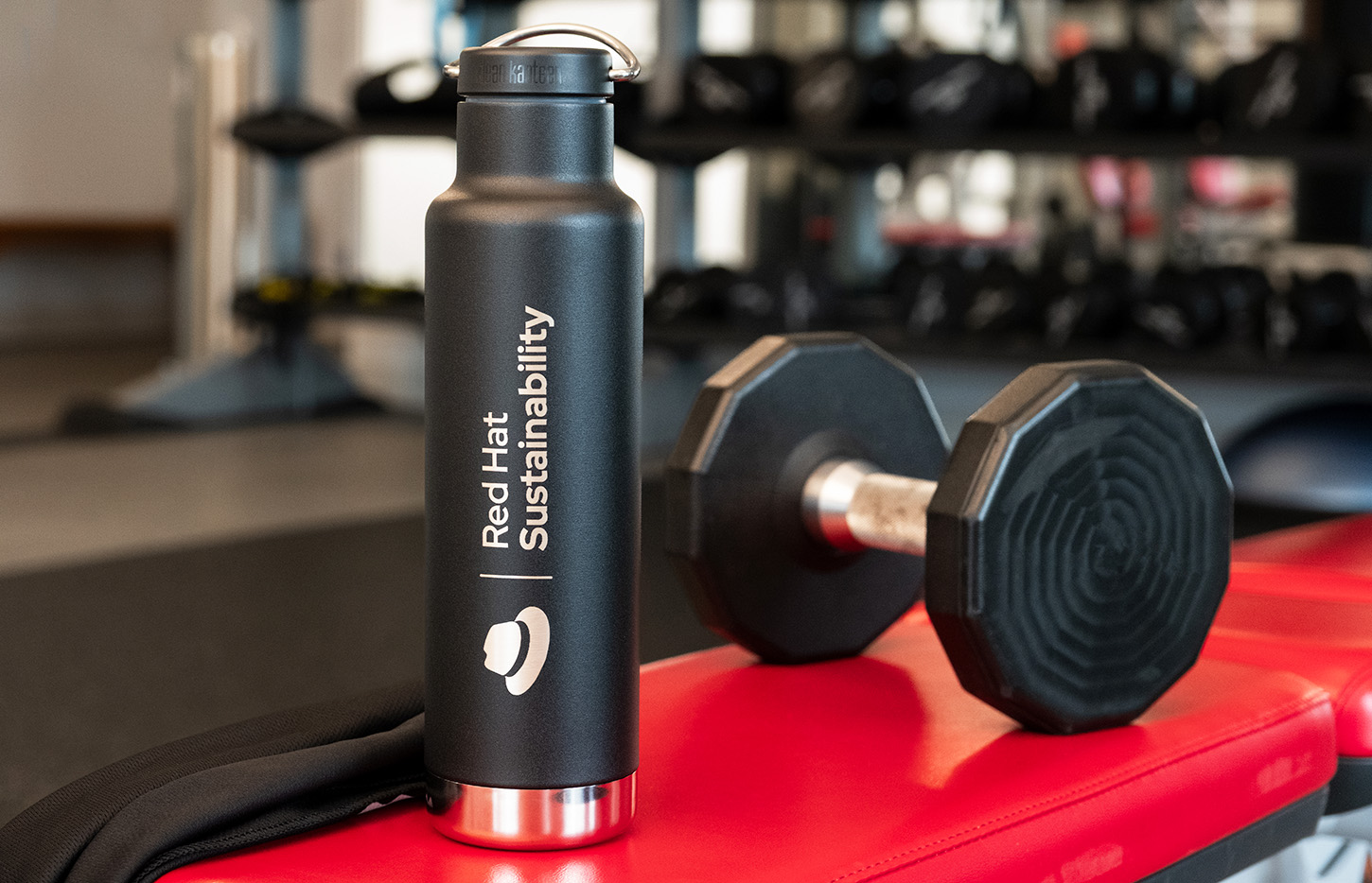 A metal waterbottle with an engraving of the Red Hat Sustainability logo, sitting on a weight bench in a gym.