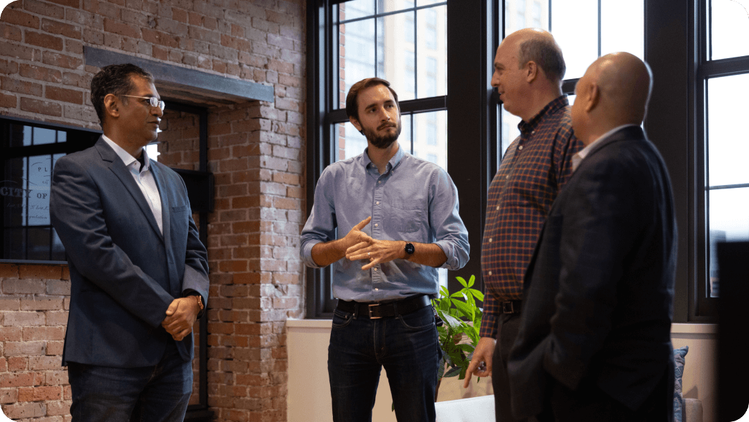 Four professionals in business casual attire talking in a modern office in front of a brick wall