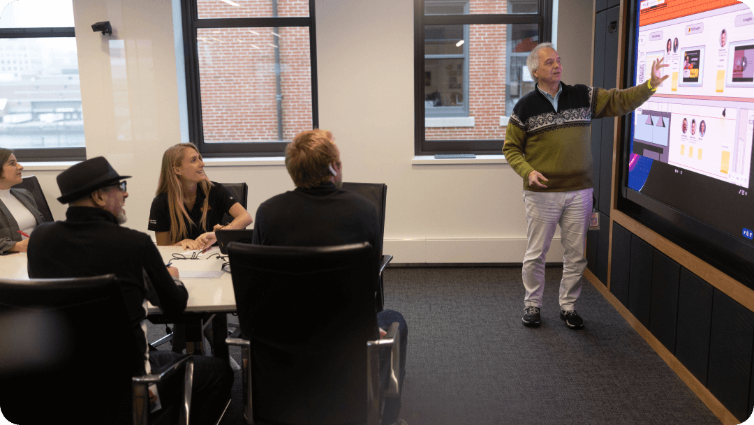 A man in a sweater presents to colleagues in a conference room, pointing to a large digital screen