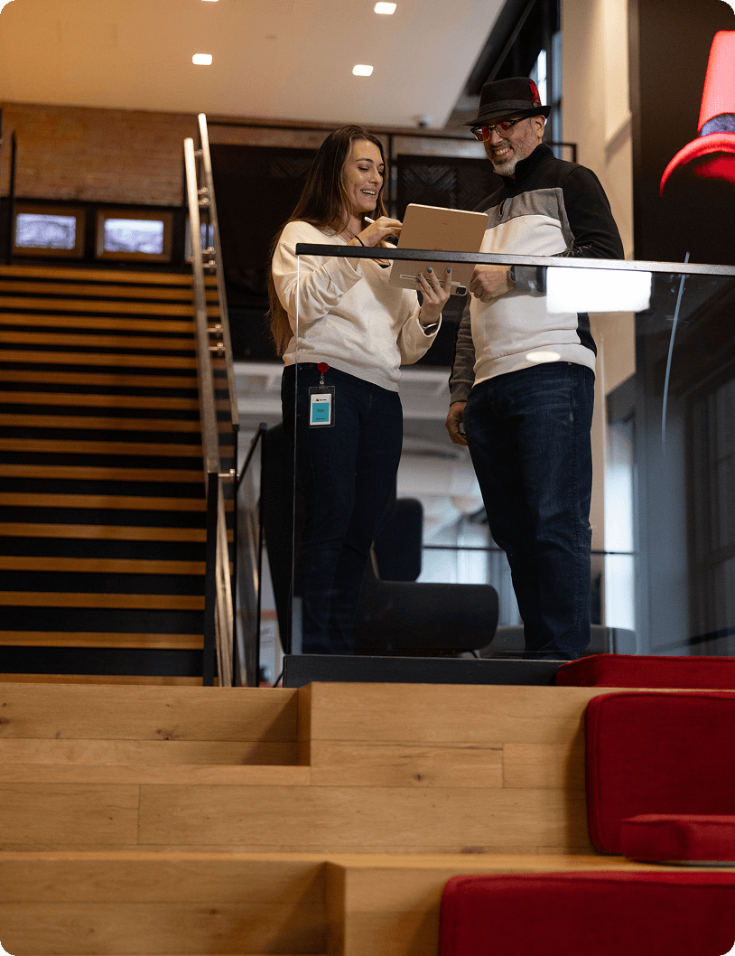 Two colleagues smile and collaborate over a tablet on a modern office staircase landing