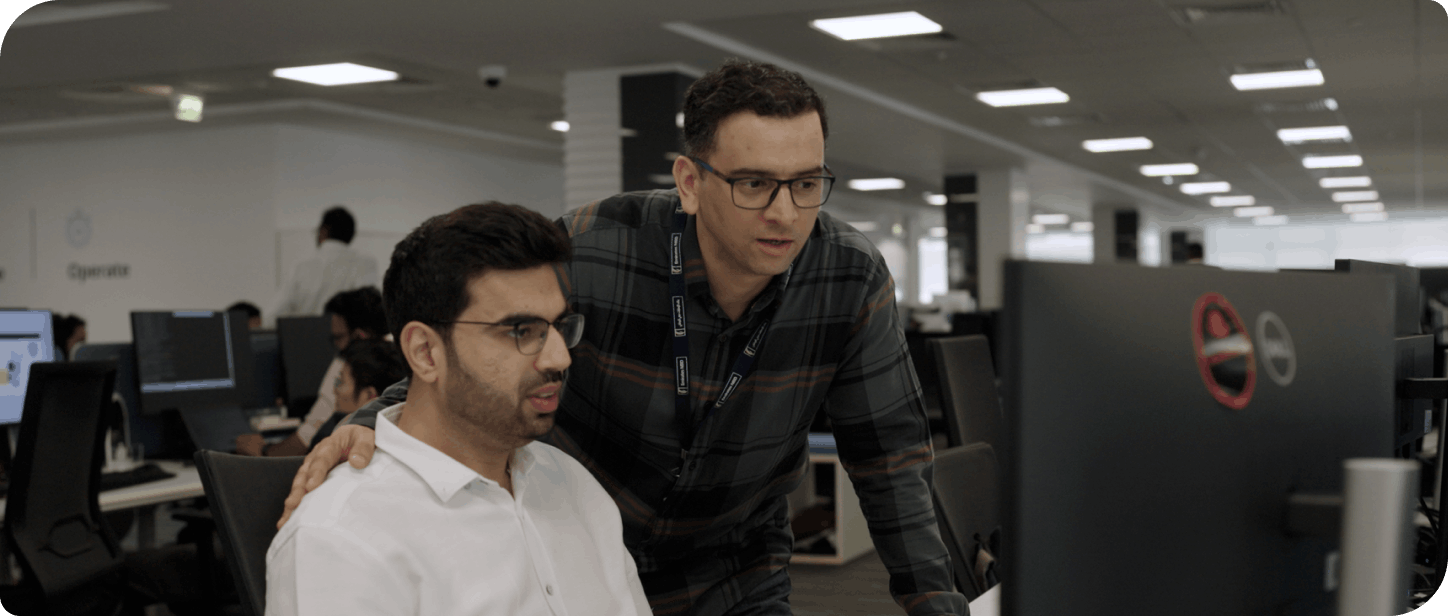 Two colleagues work together at a computer, one leaning over the other's shoulder to help