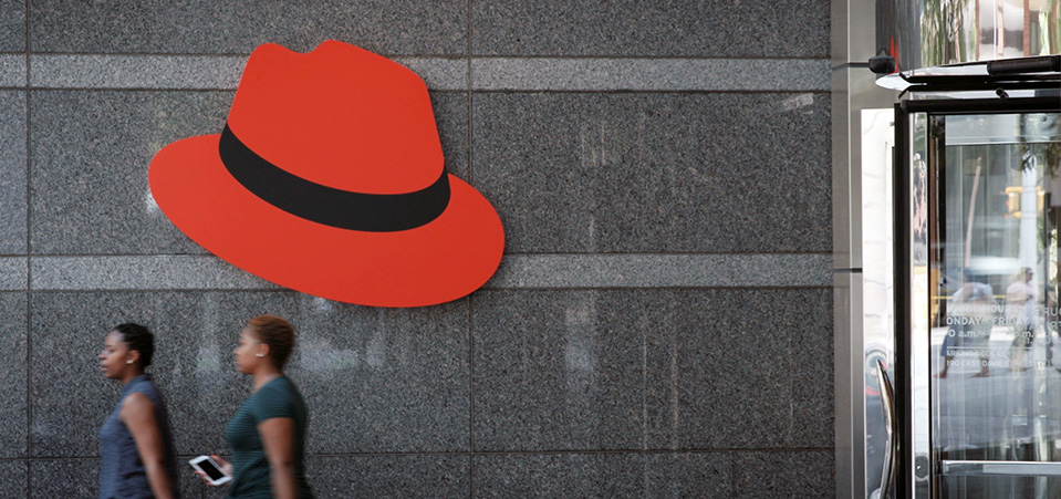 The Red Hat corporate logo on an office building exterior as two people walk by.