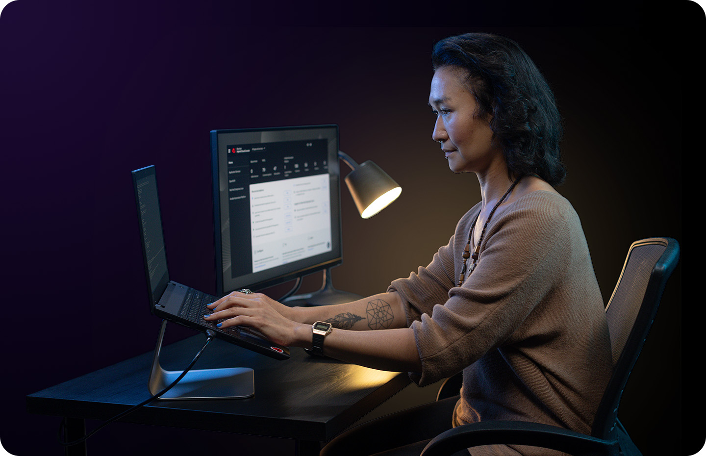 A woman focused on work at a desk with a laptop and monitor, illuminated by a lamp in the dark.