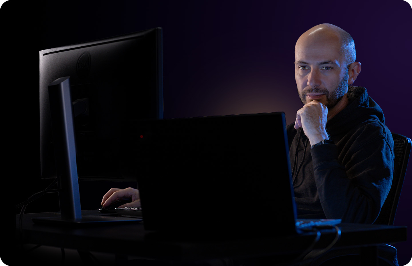 A man in a dark hoodie works at his computer in a dark room.