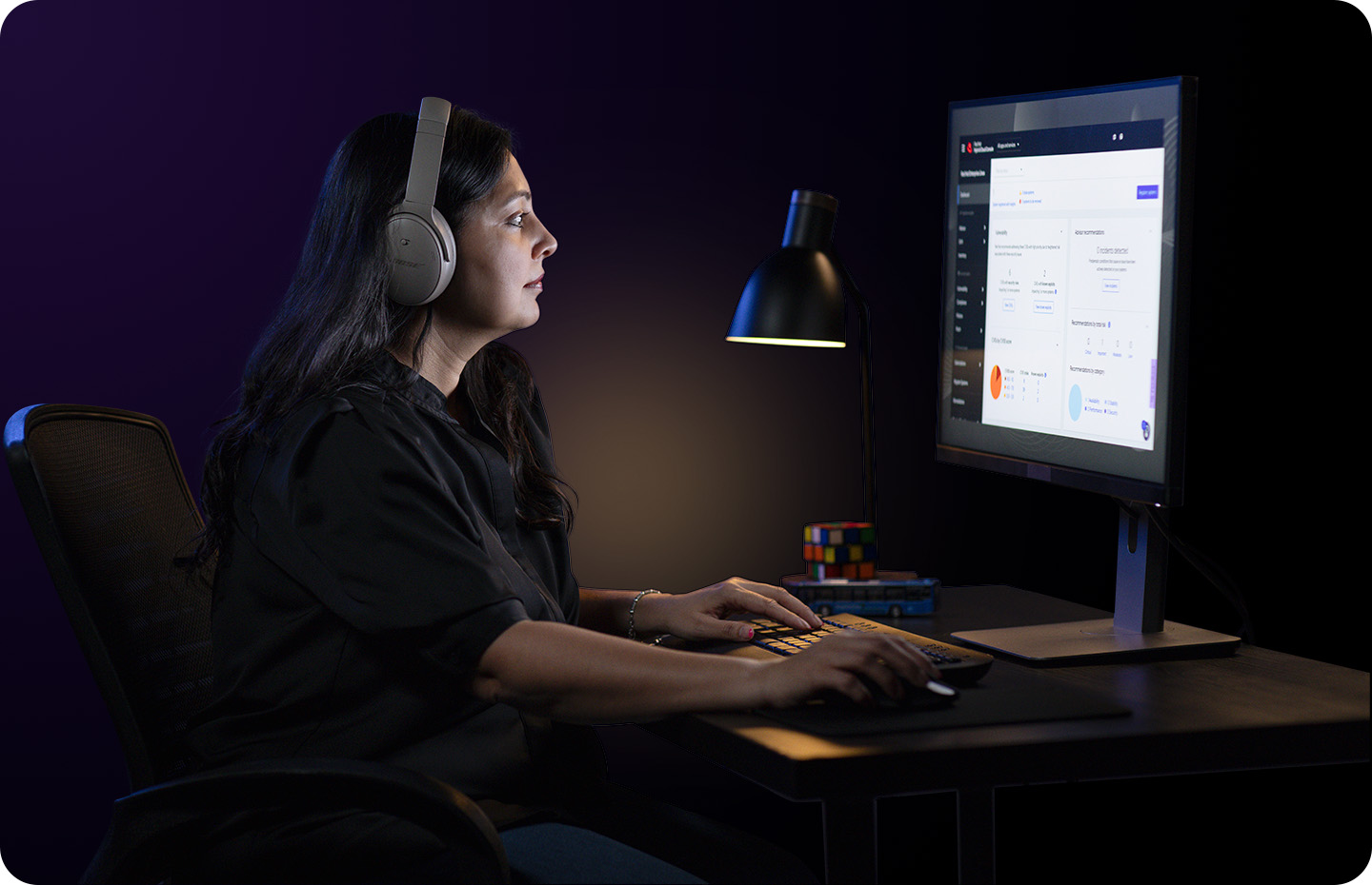 A woman in headphones works at a computer in a dark room, her face illuminated by the screen.