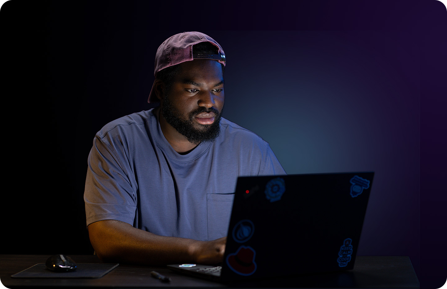 A man wearing a cap backwards works on a laptop in the dark, his face lit by the screen.