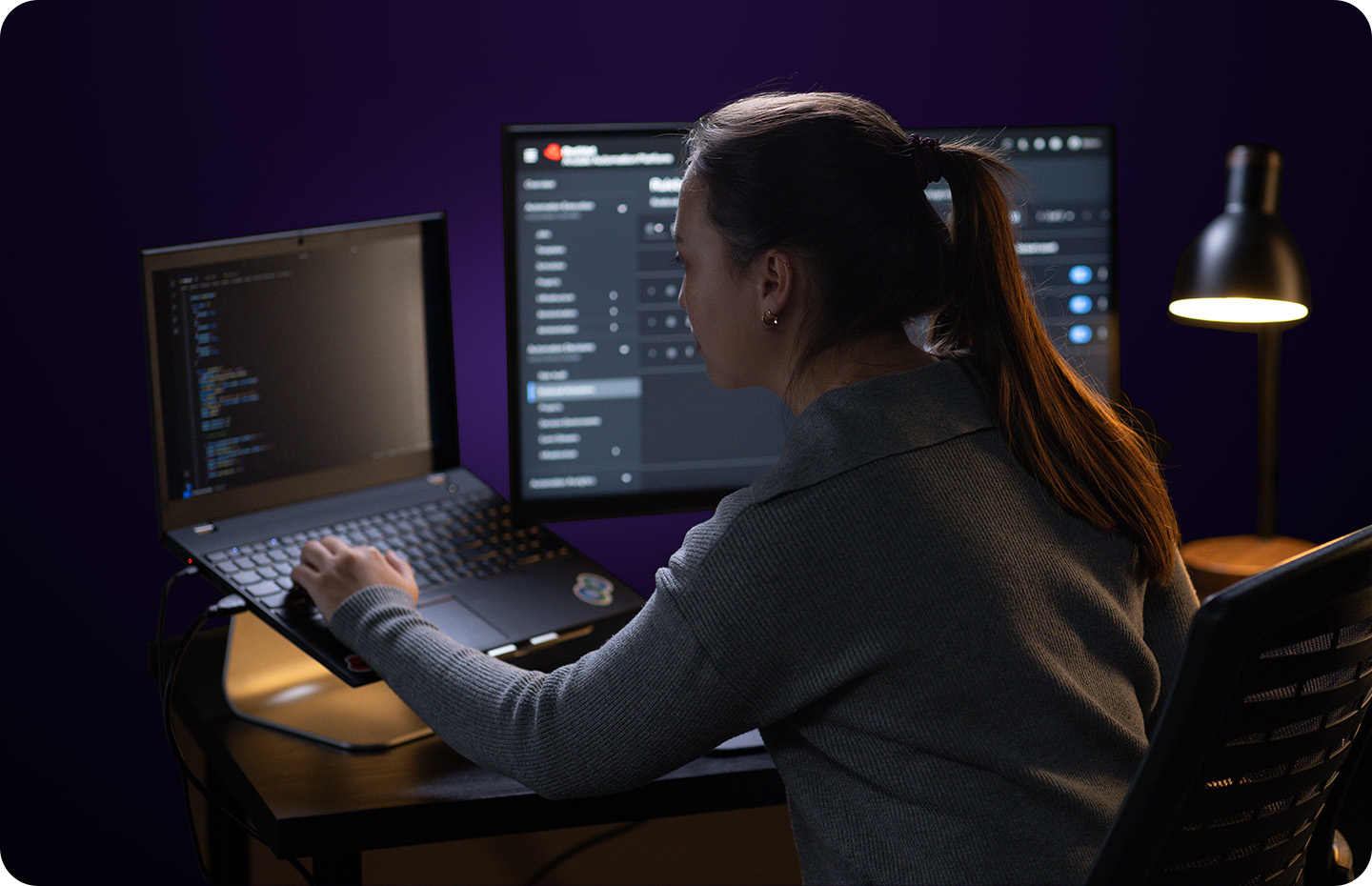 A woman at a desk with two monitors codes on a laptop in a dimly lit, purple room.