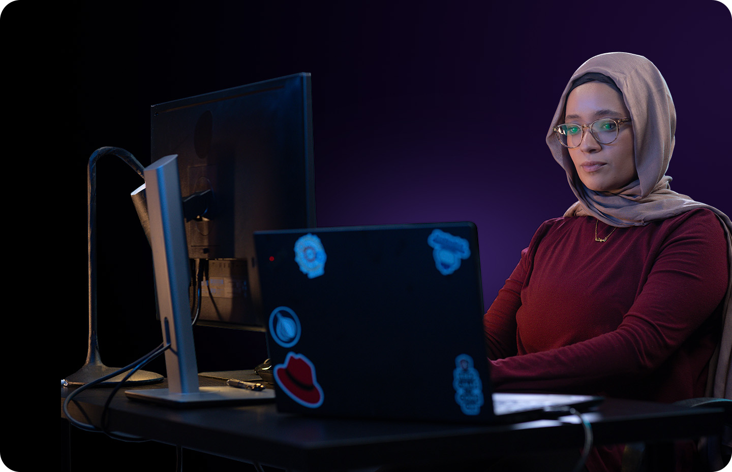 A focused woman wearing a hijab and glasses works on a laptop in a dimly lit office.