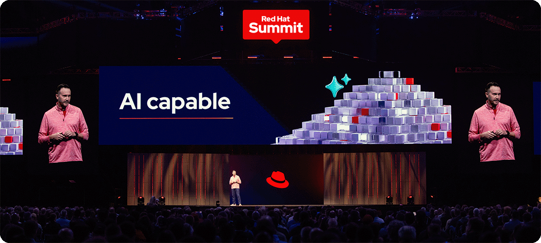 A speaker presents on stage at Red Hat Summit. The stage behind him has the Red Hat Summit logo on a screen at the top and the Red Hat fedora on a different screen at the bottom.