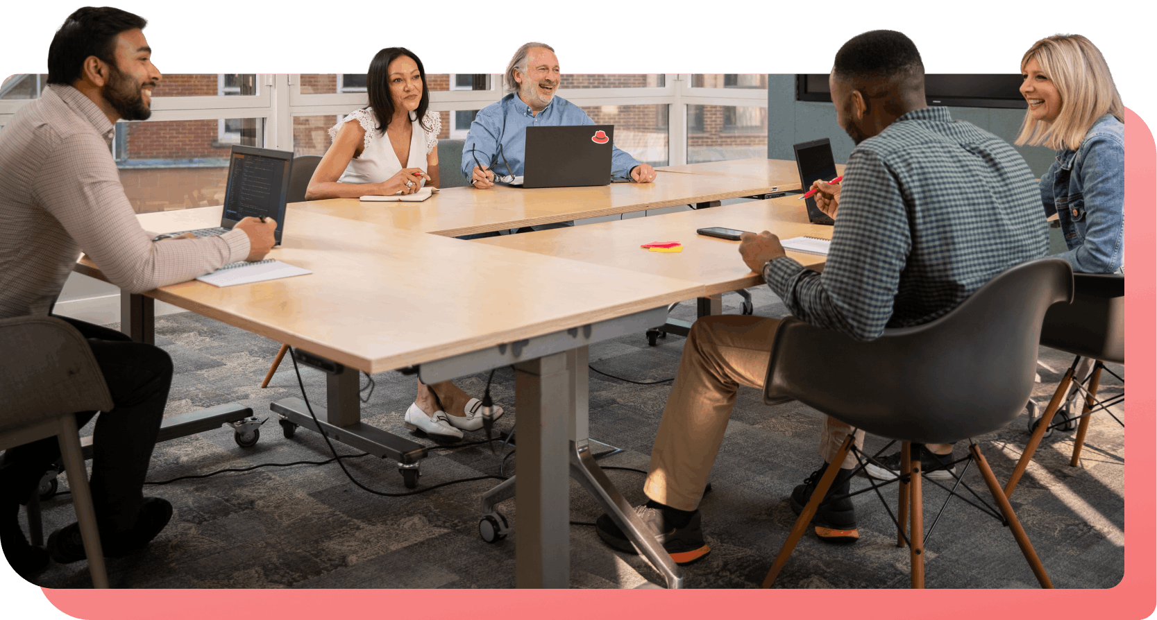 A diverse team smiles and talks during a meeting, with a laptop featuring a Red Hat logo