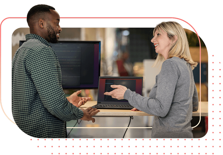 办公室内，两位面带微笑的同事在站立式办公桌前，通过一台笔记本电脑协作办公