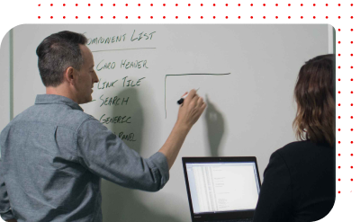A man sketches a diagram on a whiteboard as a woman with a laptop looks on