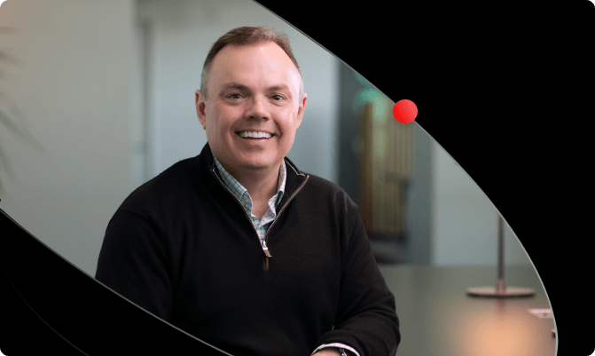 Mike Ferris in a black sweater smiles, with a black swooping graphic overlay and a small red dot