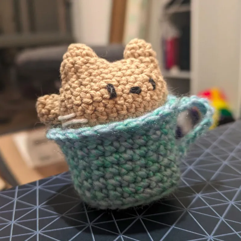 Close-up of a crocheted cotton tan-coloured cat head with a cute cartoon face, sitting in a crocheted purple-and-turquoise coffee cup