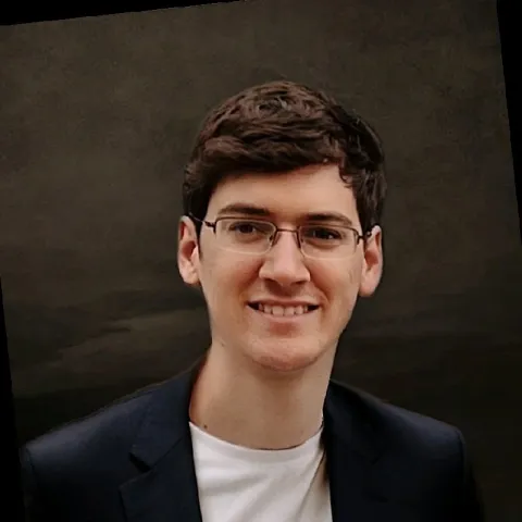Headshot of a smiling young man with brown hair, glasses, and a dark blazer