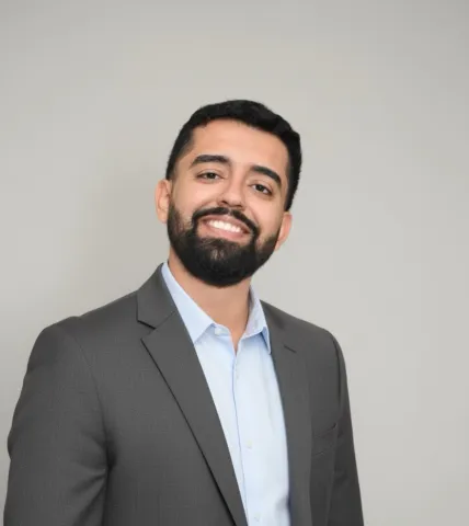 A smiling man with a beard wearing a grey suit jacket and a light blue collared shirt
