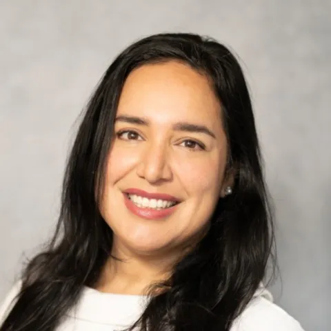 A woman named Maria Bracho with long, dark hair and a white top smiles warmly against a gray background
