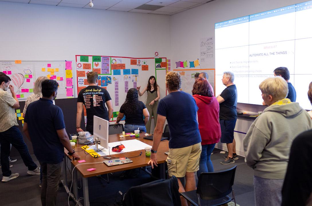 A photograph of a group of people standing around a conference table and in front of a whiteboard, continuing to brainstorm