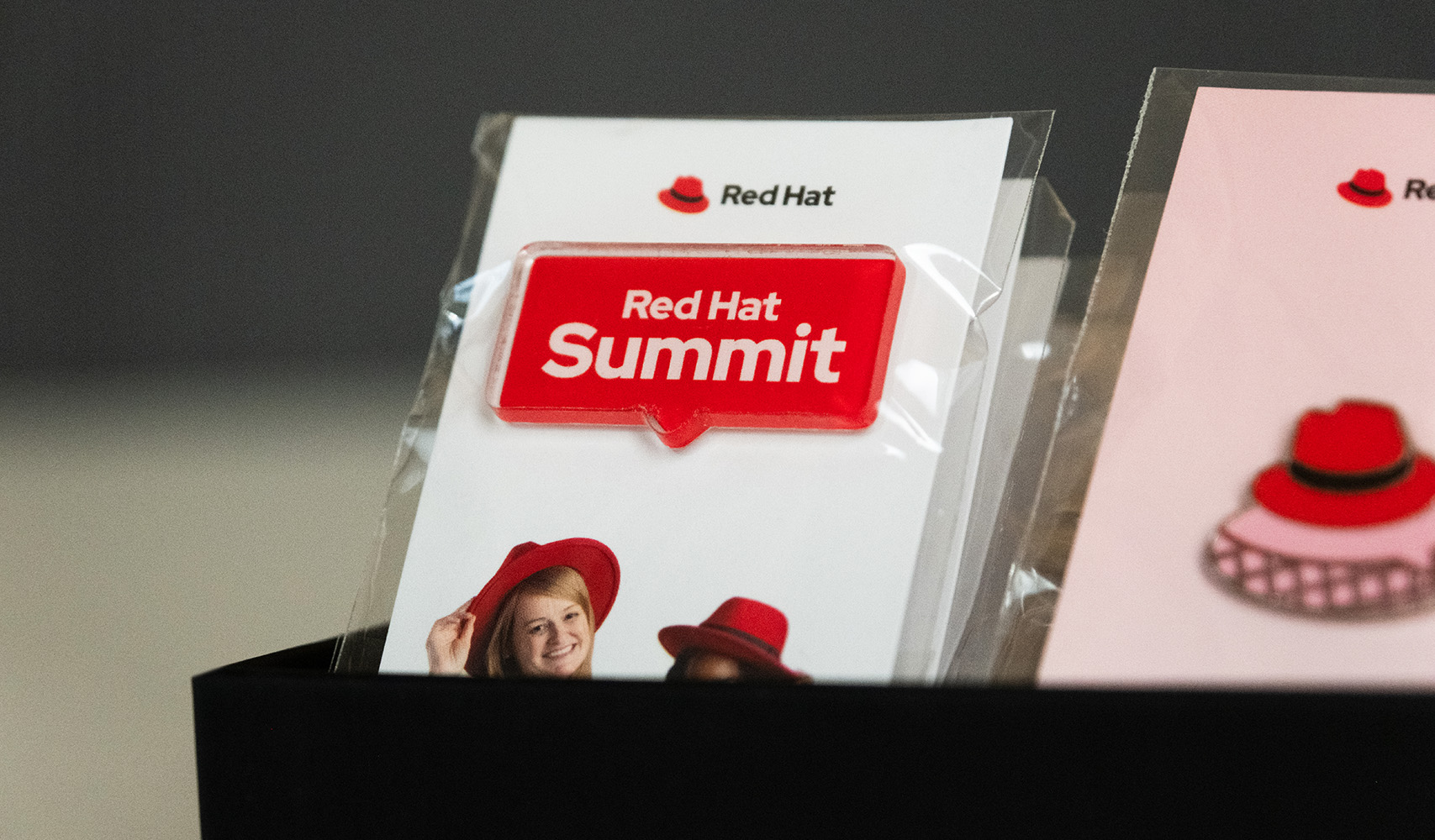 An acrylic Red Hat Summit logo pin attached to a card featuring the Red Hat logo, wrapped in cellophane.