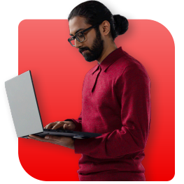 A man with a beard and glasses in a red shirt works on a laptop against a red background